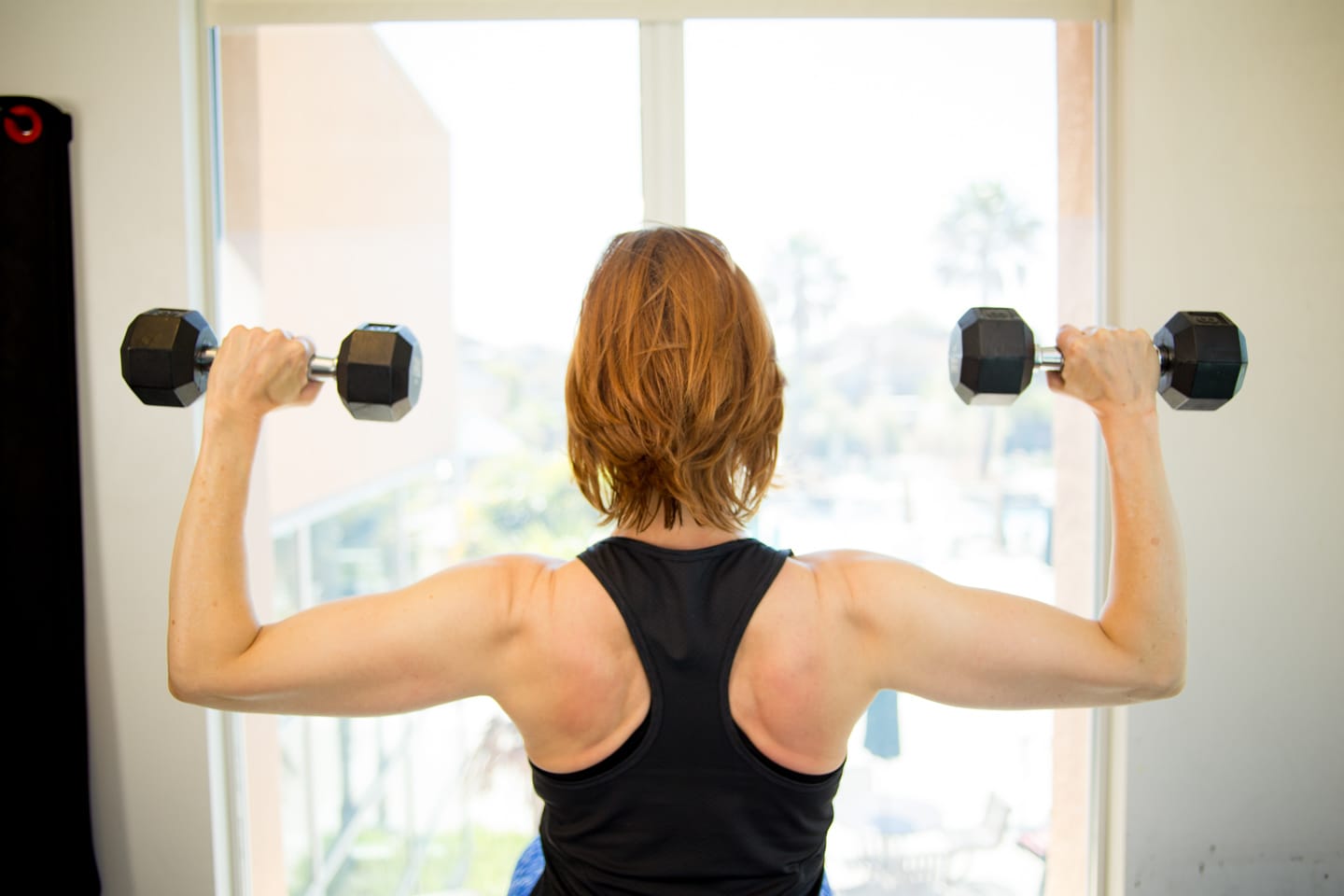 Woman indoors facing a window and lifting 8 pound weights