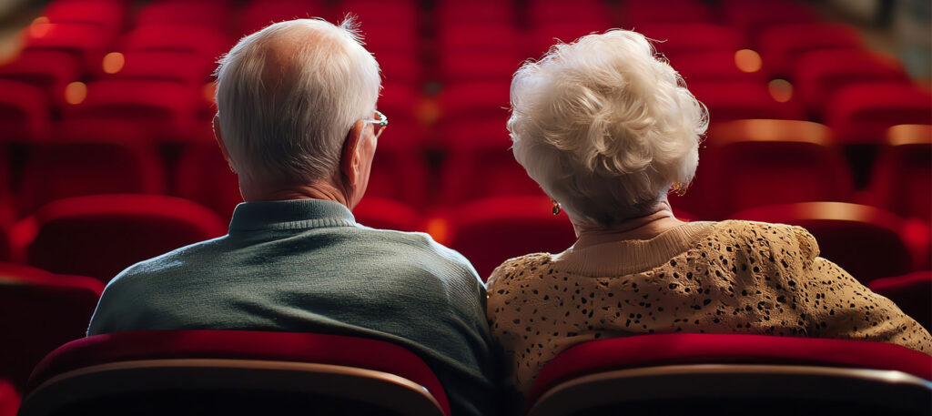 Two adults sitting together facing a theater auditorium with red velvet seats. They appear to be waiting for a live theater production to begin.